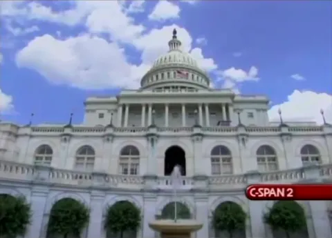 The United States Capitol building on a bright day with a blue sky and scattered clouds, viewed from a low angle; the C-SPAN 2 logo is visible in the lower right corner.