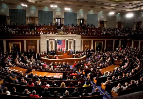 A wide-angle view of the U.S. House of Representatives chamber during a session, with members seated and an American flag displayed behind the speaker's podium.