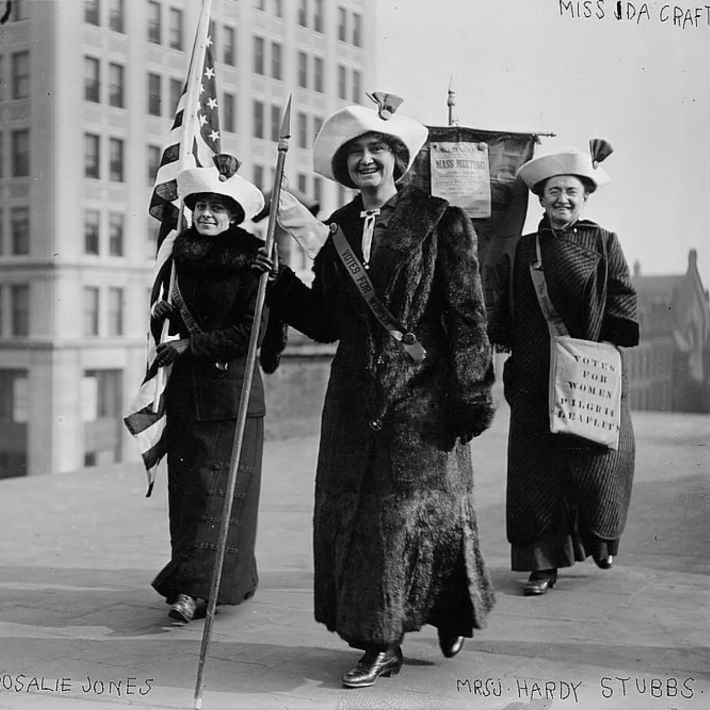 Three suffragists wearing coats and wide-brimmed hats march along a city sidewalk carrying an American flag, a staff, and a banner reading "Votes for Women."